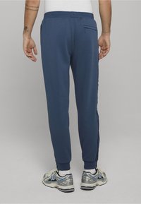 Granatowe dresy o dopasowanym kroju, z elastycznym paskiem i bocznymi paskami. Posiadają tylnią kieszeń i ściągacze przy nogawkach. Noszone z butami sportowymi.