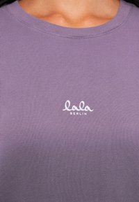 Violeta T-krekls, kas izgatavots no mīksta auduma, ar apaļu kakla izgriezumu. Baltais „lala BERLIN” logo ir novietots centrā.