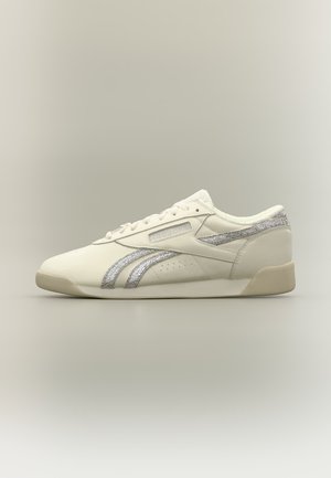 Baskets basses blanches Reebok avec bandes argentées sur les côtés, lacets à l’avant et semelle beige sur fond neutre.