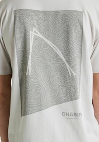 Patrón topográfico abstracto en gris y blanco con una gran línea blanca inclinada, en una camiseta gris claro con la marca "CHASIN Performance Jeanswear".