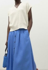 Cream knit V-neck top paired with a blue A-line skirt featuring buttons down the front and pockets; complemented by a small blue bag.