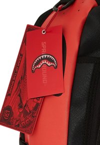 Mochila roja y negra con una superficie brillante y texturizada, con una etiqueta prominente que muestra un diseño de tiburón y el logotipo de la marca.