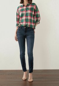 Chemise à carreaux rouge et verte à boutons avec manches retroussées, associée à un jean skinny bleu foncé et des sandales dorées à lanières sur un sol en bois.