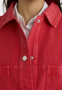 Giacca di jeans rossa con chiusura a bottoni a pressione, sopra una camicia bianca con colletto, con cuciture visibili e taschini sul petto.