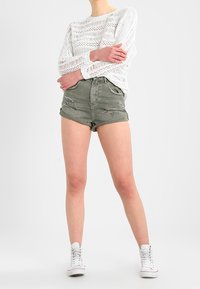 Žena v bílém pleteném topu s dlouhým rukávem, olivově zelených roztrhaných džínových šortkách a bílých vysokých teniskách, stojící se zkříženýma rukama.