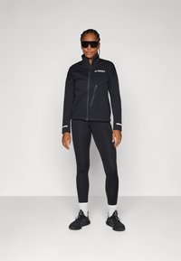 Svart Terrex-jakke med glidelås, sidelommer og reflekterende detaljer, kombinert med svarte tights og svarte joggesko.