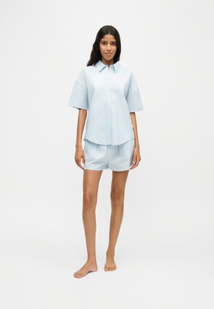 Kvinde står barfodet iført en lyseblå kortærmet skjorte med knapper og matchende shorts mod en ensfarvet hvid baggrund.