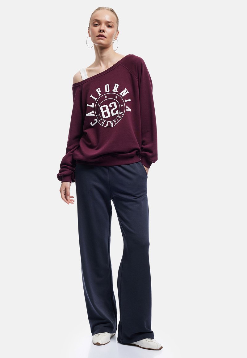 Vinröd oversize sweatshirt med vit grafisk text och logotyp. Kombineras med marinblå vida byxor och vita träningsskor. Mjuk tygtextur.