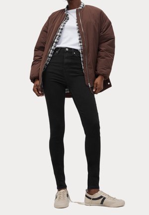 Doudoune marron sur une chemise à carreaux, t-shirt blanc, jean skinny noir et sneakers beiges avec des accents foncés. Tenue décontractée en couches.