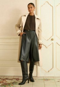 Beige esőkabát, barna plisszírozott felső, fekete műbőr rövidnadrág, térd fölé érő fekete magassarkú csizma. Mintás padló és díszes háttér előtt.