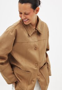 Pruun mantel kraega, esituse nupud ja kaks taskut. Omab õmblusaktsente krae ja tasku servadel. Sile tekstuur.