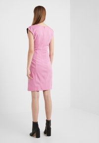 Nainen, jolla on pitkät hiukset, pukeutuneena istuvaan hihattomaan vaaleanpunaiseen mekkoon ja mustiin korkeatöppösiin, seisoo valkoista seinää vasten.