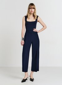 Combinaison bleu marine avec un décolleté carré et un design à jambes larges, fabriquée en tissu lisse. Associée à des mules noires avec des accents métalliques.