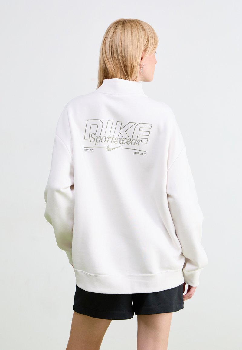 White Nike sweatshirt with high collar, oversized fit, and a large silver logo on the back; worn with black shorts. Smooth texture.