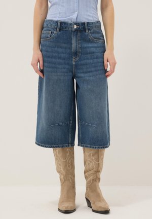 Blauwe, knielange spijkershort gedragen met beige Western-achtige laarzen met hak en een lichtblauw geruit overhemd dat in de broek is gestopt.