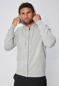 Sweat à capuche gris zippé avec une capuche à cordon, des poignets côtelés et une texture lisse. Le tissu semble doux et confortable.