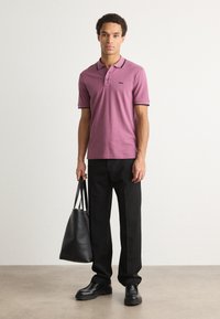 Polo majica s kratkimi rokavi plum barve z črnimi detajli, v kombinaciji s črnimi hlačami in črnimi čevlji. Manekenka drži črno torbo.