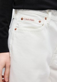 Εγγύς λήψη τσέπης λευκού τζιν Calvin Klein με χαλκινά καρφιά και ορατό μανίκι μαύρου μακρυμάνικου πουκαμίσου κοντά στο χέρι.