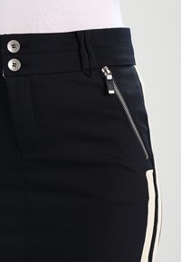 Pantalones negros con una textura suave, con herrajes plateados, un bolsillo con cremallera lateral y paneles laterales en color crema contrastante. Cintura con dos botones.