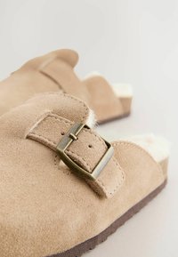 Sabots en suède beige à enfiler avec une semelle marron, dotés d'une boucle en métal et d'une douce doublure en fourrure blanche. Design plat, bout arrondi.