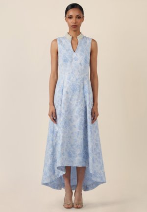 Frau stehend, trägt ein ärmelloses hellblaues Blumenkleid mit Perlenkragen und asymmetrischem Saum, kombiniert mit offenen Schuhen mit Absatz.