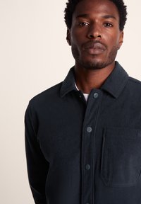 Dark blue corduroy button-up shirt with a collar, featuring vertical ribbing, a chest pocket, and round blue buttons.