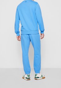 Hombre vistiendo un sudadera azul claro y un jogger a juego con zapatillas blancas y verdes, de pie contra un fondo liso claro.
