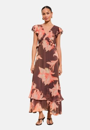 Woman wearing a brown floral wrap dress with ruffled cap sleeves and tiered hem, paired with black heeled sandals, standing against white background.