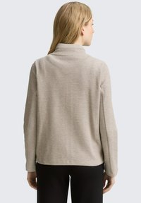 Giacca beige con texture, colletto alto, maniche lunghe e vestibilità rilassata; presenta spacchi laterali all'orlo per facilitare i movimenti.