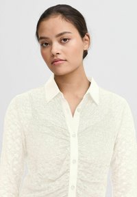Weißes Hemd mit Knopfleiste, das ein strukturiertes Spitze-Muster, einen subtilen Glanz und einen klassischen Kragen aufweist.