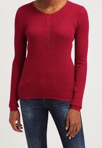 Rode geribbelde lange mouwen trui met een ronde halslijn en een vijfknopen-sluitingdetail. De stof heeft een gestructureerde en licht elastische textuur.