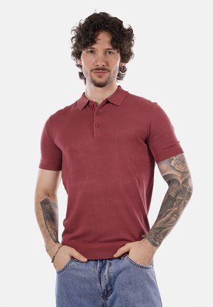 Uomo con capelli ricci, polo a maniche corte color amaranto, jeans blu e tatuaggi dettagliati su entrambi gli avambracci, in piedi con le mani in tasca.