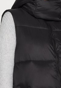 Schwarze gepolsterte Weste mit Kapuze, ausgestattet mit einer glatten Außenseite, einem Reißverschluss auf der Vorderseite und sichtbaren Nähten an einem hellgrauen Strickärmel.