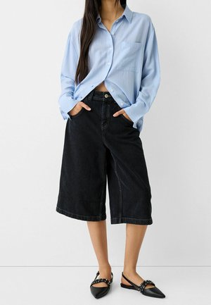 Woman wearing a light blue button-up shirt tucked into black wide-leg knee-length shorts and black pointed flats with studded straps.