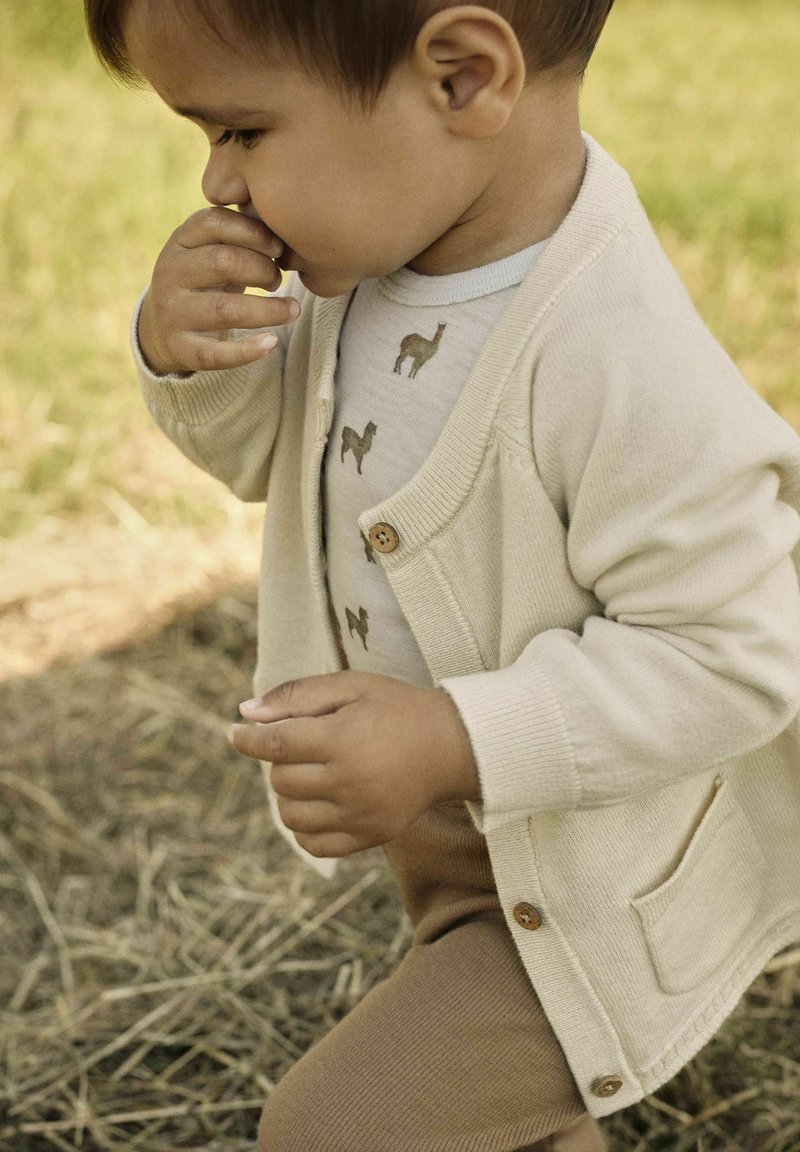 Helles beige Cardigan mit rundem Halsausschnitt, Holzknöpfen, getragen über einem cremefarbenen Hemd mit Kamelmuster, kombiniert mit braunen Ripp-Hosen.