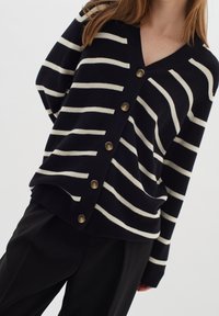 Námořnický pruhovaný svetřík se krémovými vodorovnými pruhy, výstřihem do V a šesti zlatými knoflíky vpředu. Měkká pletenina.