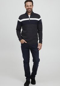 Pullover a maglia navy e grigio con striscia bianca, collo alto e zip. Indossato con jeans in denim scuro e scarpe nere, il modello si trova davanti a uno sfondo neutro.