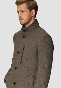 Bruine textuurjas met een hoge kraag, voorzien van zwarte knopen en een ritszak aan de linkerzijde. Glad, warm materiaal.