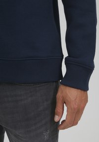 Felpa in pile blu navy con polsini e orlo a coste, indossata sopra jeans grigi strappati. Primo piano dell'area del polso che mostra la texture e la vestibilità.