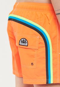 Pantaloni da bagno arancioni con strisce laterali nere, blu e verdi. Include una tasca cargo con un logo circolare sul retro.