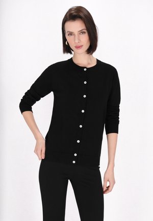 Femeie cu păr drept și închis la culoare poartă un cardigan negru cu mâneci lungi nasturat și pantaloni asortați, stând în fața unui fundal alb simplu.