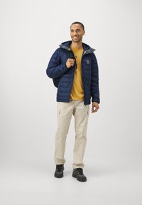 Námornícka puffová bunda so sivým vnútrom, žltými akcentmi a logom na hrudi, nosená cez žlté tričko, v kombinácii s béžovými cargo nohavicami a čiernymi topánkami.