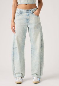 Φαρδιά τζιν παντελόνια από ανοιχτό μπλε denim με ξεβαμμένο πλύσιμο, μπροστινές τσέπες, κλείσιμο με κουμπί και ελάχιστες ραφές. Φορεμένα με λευκά αθλητικά παπούτσια.