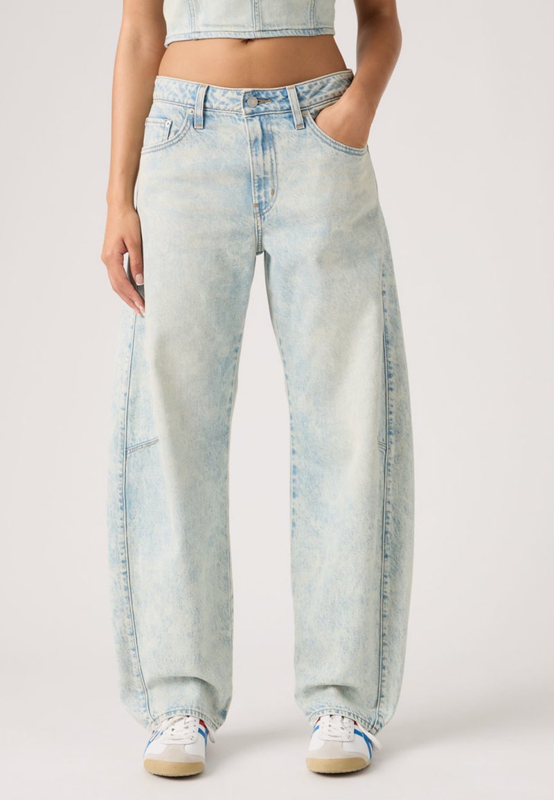 Φαρδιά τζιν παντελόνια από ανοιχτό μπλε denim με ξεβαμμένο πλύσιμο, μπροστινές τσέπες, κλείσιμο με κουμπί και ελάχιστες ραφές. Φορεμένα με λευκά αθλητικά παπούτσια.