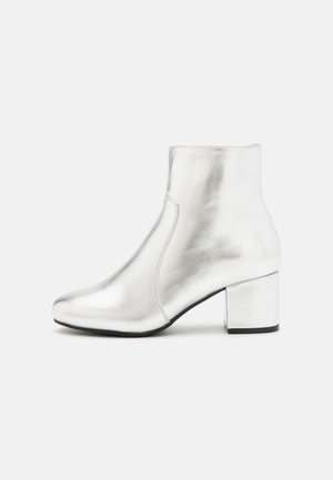 Bottine argentée en cuir brillant, avec un bout arrondi et un talon carré. Texture lisse avec des détails de coutures minimaux.