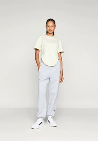 Junge Frau mit geflochtenem Haar, die ein cremefarbenes T-Shirt, hellgraue Jogginghosen und weiße Sneaker trägt, steht mit den Händen in den Taschen vor einem schlichten Hintergrund.