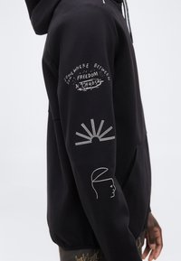 Μανίκι μαύρης μπλούζας με κουκούλα και λευκή γραμμή τέχνης: κείμενο «Κάπου ανάμεσα στην ελευθερία & το χάος», μια ακτίνα ήλιου και ένα μινιμαλιστικό προφίλ ανθρώπινου προσώπου.