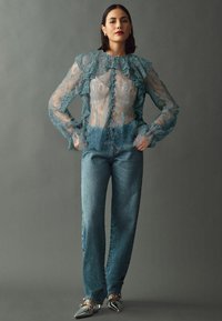Blouse en dentelle turquoise transparente avec un col et des poignets volants, associée à un jean flare bleu clair et des chaussures métalliques à bout pointu.