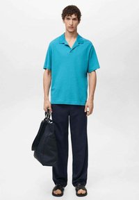 Tirkizna polo majica s kratkimi rokavi, ohlapne črne hlače, črne sandale in temna torba. Gladka tekstura, preprost dizajn, brez vidnih vzorcev.