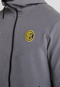 Sweat à capuche gris avec fermeture éclair, fabriqué en tissu doux, arborant un logo circulaire jaune et une fermeture éclair noire. Texture lisse avec des détails de coutures.